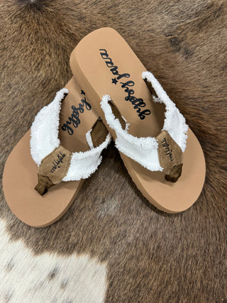 A pair of beige Snow Flip Flops with white fabric straps and brown accents are placed on an animal hide rug. These comfortable flip flops from Western Edge Boutique have a casual, beachy look, with "Western Edge Boutique" branding visible on the footbed and fabric straps.