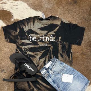 Make a bold statement with the hand-bleached black and brown tie-dye "BE KIND" T-shirt from Western Edge Boutique. It features the striking phrase "be kind...of a bitch" on its unique design, perfectly complemented by denim shorts with a tag and black flip-flops showcasing elegant cursive writing, all displayed on a brown and white cowhide rug.