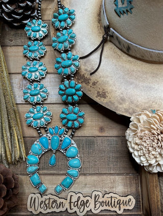 The Arizona Squash Blossom Necklace, radiating beauty with its turquoise stones and intricate silver accents, is showcased on a rustic wooden background. Nearby, a beige cowboy hat embellished with blue embroidery sits alongside a faux flower and pinecones. A "Western Edge Boutique" sign at the bottom completes this stunning display.