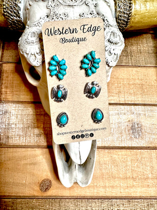 A display card from Western Edge Boutique showcases three pairs of Cylan turquoise-themed stud earrings with burnished silver details. The card is pinned to a decorative skull accented with lace, set against a rustic wooden background. The lower part of the card features the boutique's website and social media icons.
