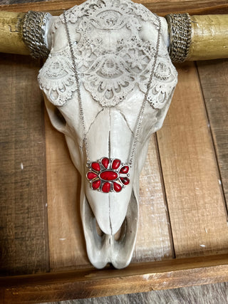 A decorated animal skull with ornate lace patterns and golden horns is mounted on a wooden background. A Carolina Pendant Necklace by Western Edge Boutique, featuring red-colored stones and a 14″+3″ extension, is draped over the skull's forehead.