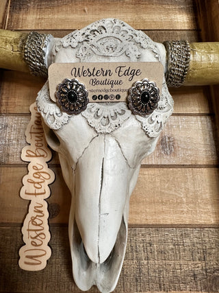 A decorative cow skull with horns is displayed against a wooden backdrop. Adorned with burnished silver jewelry and a business card for "Western Edge Boutique" placed on its forehead, it showcases the rustic elegance of Delilah Black Opal Concho Stud Earrings. A wooden cutout with the same business name is attached to one horn.
