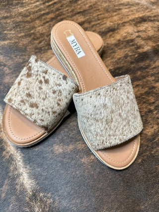 A pair of Baylee Cowhide Leather Flats by Myra, featuring light brown and white cowhide straps adorned with golden rivets, are placed on a surface that resembles cowhide. These sandals showcase a beige sole and have a tag with text on the inner lining.