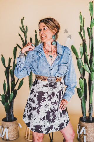 A woman poses confidently in front of a beige background with potted cacti. She wears the Mackey Western Chambray and Cow Print Dress by 2 Fly, accented with turquoise jewelry including earrings and a necklace. Butterflies appear to her right.