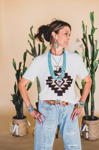 A woman stands confidently with her hands on her hips, wearing the Tangee Teca Taos Tee from 2 Fly, which features a geometric pattern. She pairs it with ripped blue jeans made from a comfortable spandex blend and layers turquoise necklaces. Her look is further accessorized with matching earrings, bracelets, and a belt. Potted cactus plants are visible in the background.