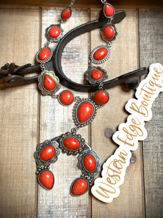 The Blaney Chunky Squash Necklace from Western Edge Boutique, featuring vivid red stones set in burnished silver, is artistically displayed on a wooden surface. The ensemble includes a necklace with a striking pendant and a pair of matching earrings. A wooden tag engraved with "Western Edge Boutique" rests to the side.