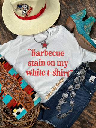 A flat lay display featuring the super soft "BBQ Stain on my White T-Shirt Graphic Tee" from Western Edge Boutique, a fringed turquoise and brown bag, blue jeans, turquoise cowboy boots, a western hat with red accents, a star necklace, a silver belt, and a woven bracelet. Ideal for summer BBQ parties!