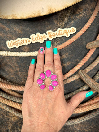 A hand with turquoise-painted nails and a small cactus design on the ring finger is wearing the Colby Western Squash Stretch Ring, which features a large, pink, oval gemstone set in burnished silver. The background showcases rope and a "Western Edge Boutique" sign in cursive.