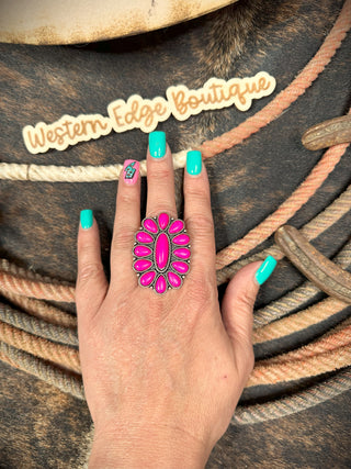 A hand with teal nail polish and a lightning bolt design on one nail displays the Starla Western Conch Cuff Ring, featuring large fuchsia color stones and smaller ovals set on a burnished silver band. The background showcases rustic ropes and a "Western Edge Boutique" sign.