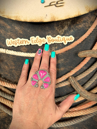 A hand with turquoise nail polish and a pink design on the ring finger, adorned with the Brynna Western Squash Stretch Ring in large burnished silver and pink, is placed on coiled ropes. A "Western Edge Boutique" sign is in the background.