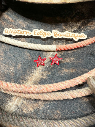 A pair of Star Stone Stud Earrings, featuring a coral star shape and adorned with tiny Aurora Borealis stones, rests on a textured brown and beige surface. Above the earrings is a sign from Western Edge Boutique, decorated with a rope-like design.