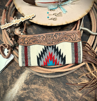 A handcrafted Cameron Saddle Blanket and Tooled Leather Clutch by American Darling, made from Genuine Full Grain Leather and featuring intricate Native American-inspired patterns, is placed on top of a coiled lasso. A feather-decorated cowboy hat with turquoise beads sits in the background alongside a sign reading "Western Edge Boutique.