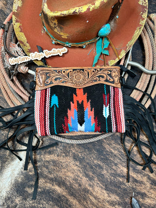 The Braxlyn Saddle Blanket and Tooled Leather Handbag by American Darling is showcased against a rustic background, highlighting its handcrafted design and intricate detailing with a vibrant tribal pattern in red, blue, and white. Made from hand-tooled vegetable tanned cowhide leather, the handbag features black fringe accents and an adjustable, detachable shoulder strap. Beside it rests a brown wide-brimmed hat adorned with turquoise and a feather.