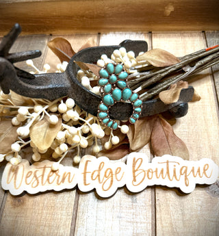 A rustic display featuring a pair of turquoise earrings set on a decorative metal holder with dried floral elements. Below the earrings, alongside the exquisite Colette Squash Adjustable Ring, there is a sign that reads, "Western Edge Boutique." The background has a wooden texture.