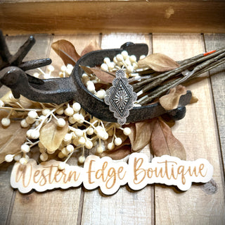 An assortment of decorative items sits on a rustic wooden surface. Highlighted is an antique silver, diamond-shaped brooch with intricate detailing, nestled among beige leaves and white berries. In the foreground, a sign reads "Western Edge Boutique" in a stylish cursive font, while the elegant Brigen Boho Concho Adjustable Ring with Double Band exudes boho charm.