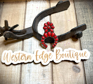 An intricately designed Colette Squash Adjustable Ring in a vibrant red Western style is nestled within an old cowboy spur. Below the ring, a wood-cut sign elegantly reads "Western Edge Boutique" in cursive script. The display rests on a rustic wooden surface.
