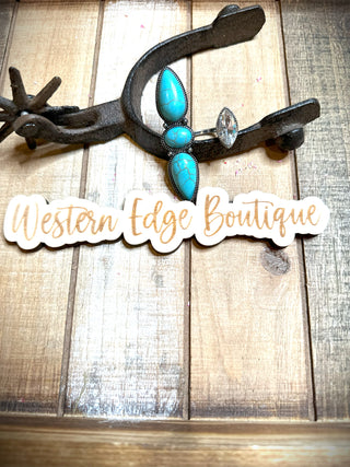A rustic wooden background showcases a metal horseshoe and spur. Below them, turquoise jewelry and the stunning Trixie Turquoise Cuff Ring glimmer in the light. The text "Western Edge Boutique" is written in a stylish script on a white cutout.