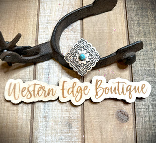 Placed on a rustic wooden backdrop, the Amelia Concho Adjustable Ring from Western Edge Boutique showcases a decorative Western-style antique silver ring with a turquoise stone, resting on an old iron tool. Below the tool, an elegantly designed wooden cutout sign displays "Western Edge Boutique" in a cursive font.
