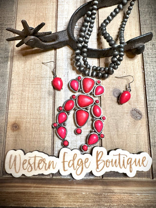 A vibrant red and burnished silver Sunny Navajo Pearl Squash Necklace Set with matching earrings hangs on a rustic metal display against a wooden background. The necklace dazzles with large, red color stones outlined in silver. Below the jewelry is a sign that reads "Western Edge Boutique.