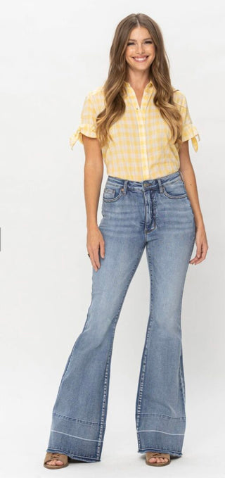A woman with long, wavy hair smiles while standing against a white background. She is wearing a short-sleeved yellow checkered blouse and blue Townes High Waisted Tummy Control Judy Blue Flares from Western Edge Boutique, paired with tan platform sandals.