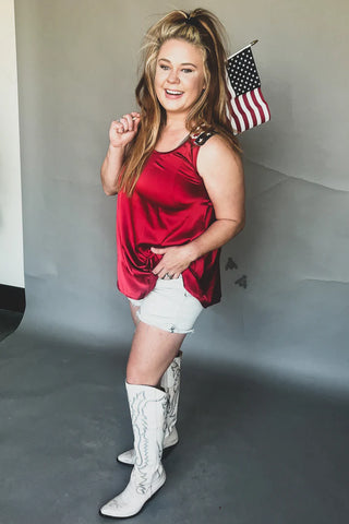 A woman stands smiling, wearing THE BETSY ROSS red tank top with embroidered straps by 2FLY, along with white shorts and white knee-high boots featuring a wavy pattern. She has long blonde hair styled in a high ponytail, with an American flag serving as the backdrop.