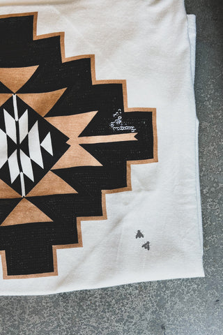 The Tayten Teca Taos 2.0 by 2 Fly is a super soft white fabric adorned with a graphic print of black and tan geometric patterns, showcasing a large central diamond surrounded by zigzag shapes. Adding a unique touch, two tiny motifs resembling insects or bees are embroidered near the edge of the speckled gray background.