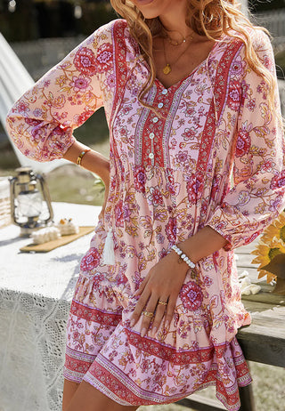 A woman is wearing the Avery Pink Floral Boho Print dress by Western Edge Boutique, featuring a tassel tie neckline. She sits outdoors near a table decorated with a lace tablecloth and small lantern. Her accessories include rings and a bracelet, while the grassy background is illuminated by sunlight.