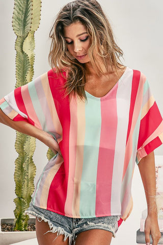 A woman with tousled hair stands near a tall cactus, wearing an Iris Multi Stripe Summer Vibes Blouse by BiBi in vibrant colors. The blouse features a colorful, vertically striped design with a V-neckline and is paired with denim shorts. She looks down, one arm resting on her hip against the minimal and neutral background.