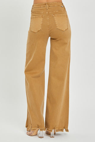 The image showcases a person from the waist down wearing Caliber Camel High Rise Wide Pants by Risen, characterized by their high-waisted fit and brown wide-leg design with raw, frayed hems. These stylish jeans feature back pockets and are complemented by clear heeled shoes against a plain, light-colored background.