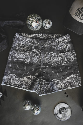 SHE GON' COUNTRY mesh shorts from 2 Fly, featuring an intricate black and white pattern with various text and images, are placed on a dark surface surrounded by small disco balls and a sequined hat. The striking design of these shorts adds a visually captivating contrast to the surrounding items – perfect for summer events.