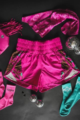 The 2 Fly Azyee Shorts Fushia, featuring a vibrant pink hue and colorful Southwestern pattern made from athletic fabric, are displayed on a black background. Surrounding the shorts are a matching pink bandana, pink socks with "YEEHAW" printed on them, a pink fringe accessory, teal socks, and small disco balls.
