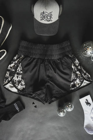 A flat lay featuring the "Short Round Midnight" black athletic shorts from 2 Fly with a white pattern on the sides, made from athletic fabric, accompanied by a matching black cap with a white emblem, black socks, white socks with a black logo, disco balls, and small black decorative items on a dark surface.