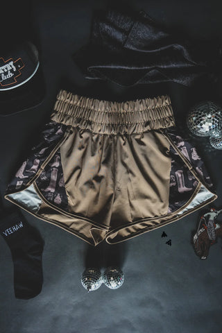 A pair of 2 Fly's Short Rounds Saddle, made from beige athletic fabric and featuring a ruched waistband with cute guitar print accents, is laid flat on a dark surface. The shorts are surrounded by items including a black baseball cap, black socks with text, small disco balls, a keychain, and other accessories.