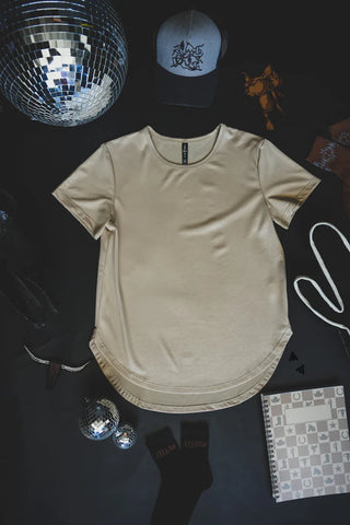 A beige t-shirt, specifically the Swanky Faux Leather Cotton Shirt by 2 Fly, is laid out on a black surface surrounded by various items: two disco balls, a gray cap, a scarf, a patterned notebook, black socks, sunglasses, a hanger, and intricately designed clothing pieces. The setup exudes a stylish and eclectic vibe with rad leather lux touches adding an edgy flair.