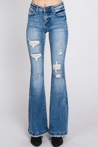 A person wearing Petra153's Audrey Distressed Mid Rise Stretch Flare Jeans with distressed details on the thighs and knees stands against a white background. Featuring 5-pocket styling and a button and zipper closure, the jeans' hem slightly covers black shoes, blending modern fashion with classic design.