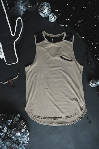 A light brown Dolly Tank DUST by 2 Fly, featuring a zippered chest pocket and dark shoulder patches, is laid out on a dark surface. Surrounding it are various silver and black decorations, including a disco ball, sequins, and a cactus ornament, evoking retro rodeo vibes.
