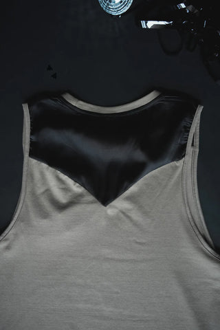 A muscle style tank called the "Dolly Tank DUST" by 2 Fly, featuring a beige body with a black satin-like V-shaped design on the back, is laid on a dark surface accompanied by scattered small dark objects and a piece of reflective material partially visible at the top.