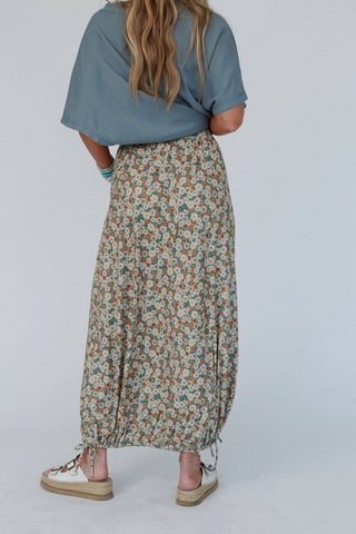 A person stands with their back to the camera, donning a light blue top paired with the Solstice Song Cinched Skirt in khaki by Three Bird Nest. They have long, wavy hair and are sporting white sneakers. The skirt features an adjustable drawstring hem that provides a cinched look, contributing to a casual and summery overall style.