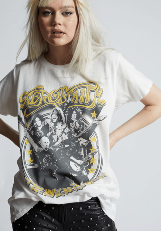 A person with long blonde hair is wearing a white vintage Emotion 80s Rock Band Aerosmith Unisex Tee by Recycled Karma, featuring images of the band members and the text "Let The Music Do The Talking." They are also wearing studded black pants and standing against a plain background, exuding 80s rock nostalgia.