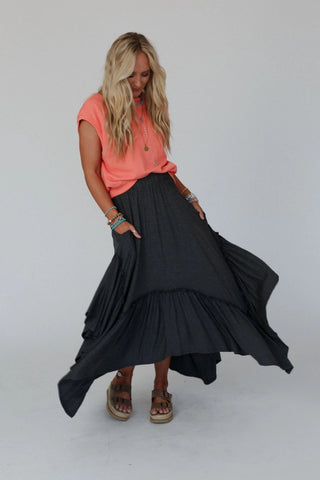 A woman with long blonde hair, wearing an orange top and the Logan Forever Beauty Maxi Skirt in a pale sage hue by Three Bird Nest, poses against a plain white backdrop. She accessorizes with bracelets on both wrists and tan sandals. Gazing downward, she holds out the lightweight stretch knit fabric of her high-rise boho maxi skirt.