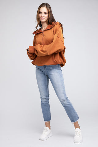 Wearing a Burke Quarter Zip Stitch Detailed Elastic Hem Hoodie from Western Edge Boutique in brown and pairing it with light blue jeans, a person exudes fashion confidence. They pose against a plain gray background with hands slightly crossed, completing the look with white sneakers.