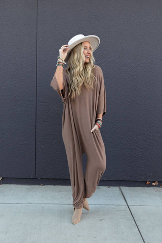 A person with long, wavy blonde hair stands against a dark gray wall. They are wearing the Lovely As Ever Harem Jumpsuit by Three Bird Nest, made of comfortable knit fabric, and a tan hat paired with several bracelets on their wrist. They are holding the brim of the hat with one hand, smiling, and looking to the side.