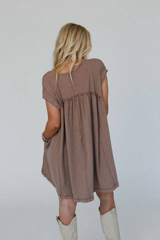 A person with long blonde hair is standing with their back to the camera, wearing a loose, short-sleeved taupe dress with a gathered waist and tall white boots. The Meadow Subtle Escape Textured Dress by Three Bird Nest, combined with boho western vibes, creates a relaxed yet stylish look. Their hands rest casually at their sides.