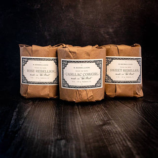 Three brown paper-wrapped soap bars with vintage-style labels that read, from left to right: "Rose Rebellion," "Cadillac Cowgirl," and "Sweet Rebellion." They are placed on a dark, textured surface against a dark background, reminiscent of the handcrafted elegance of R. Rebellion's Summer Nights Candle 8 oz., known for its wood wick charm.