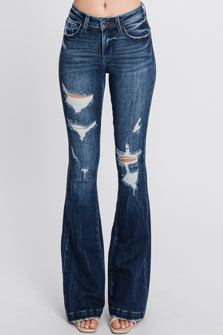 A person wearing Chevelle Distressed Mid Rise Stretch Flare Jeans by Petra153 with distressed detailing and ripped patches on both thighs. Featuring classic 5-pocket styling, the jeans are paired with open-toed shoes against a plain white background.