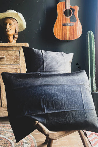 A rustic room features a wooden dresser with a bust sculpture wearing a cowboy hat. Two 2 Fly SATIN SLEEP *BOOT STITCH pillows, known for their satin-esque sheen, are stacked on a chair in the foreground. A wooden acoustic guitar and a tall cactus are mounted on a dark wall in the background.
