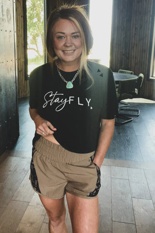 A smiling woman with blonde hair stands indoors wearing a 2 Fly black T-shirt featuring the text "Stay Fly," tan Short Rounds Saddle athletic fabric shorts, and a turquoise necklace. She has one hand in her pocket, and there is a table with chairs in the background.