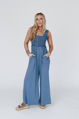 A woman with long blonde hair stands confidently against a plain white background. She is wearing the Three Bird Nest "Nothing Compares Smocked Ruffle Jumpsuit" in Denim Blue, made of lightweight stretch fabric and featuring wide pant legs and pockets. She accessorizes with layered bracelets and necklaces, wears sandals, and has her hands in her pockets.