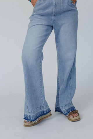 A person is seen from the waist down wearing the Cali Dream Denim Overalls in a light wash by Three Bird Nest, featuring wide legs and frayed hems at the bottom. The comfortable fabric adds to the relaxed vibe, paired with brown flip-flops that have a single strap across the toes. Their hands are tucked into the pockets of the overalls on a white background.