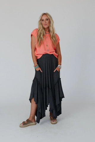 A woman with long blonde hair wears a coral short-sleeve blouse and the Logan Forever Beauty Maxi Skirt in Pale Sage by Three Bird Nest, featuring a high-rise elastic waistband. She accessorizes with multiple bracelets and necklaces, standing against a plain white background, one hand in her skirt pocket and the other resting by her side.